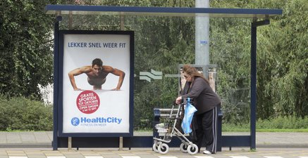 Ältere Frau mit Rollator an einer Haltestelle. Darin eine Werbung, die einen sehr starken sportlichen Mann zeigt. 
