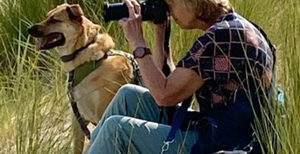 Eine Frau sitzt auf dem Sandstrand und schaut durch den Sucher einer Fotokamera. Neben ihr sitzt ein Hund.
