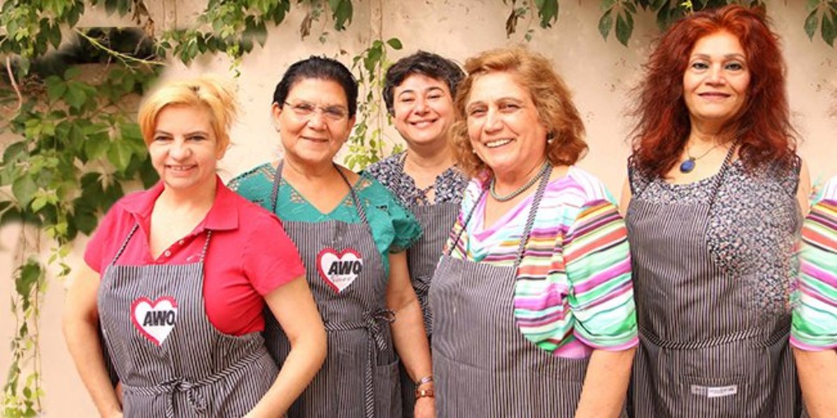Fünf ältere Frauen unterschiedlicher Herkunft mit Kochschürzen stehen vor einer Mauer