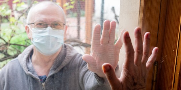 Mann mit Mund- Nasenschutz steht draußen vor einem Fenster und hält seine Hand an die Scheibe. Von innen hält eine zweite Person seine Hand gegen seine Hand. 