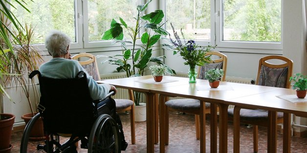 Alte Frau im Rollstuhl sitzt allein in einem Aufenthaltsraum und schaut aus dem Fenster