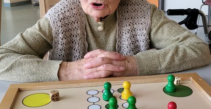 Frau in hohem Alter spielt Brettspiel. Gesicht in Falten gezogen, ruft "Mensch ärgere dich nicht!"