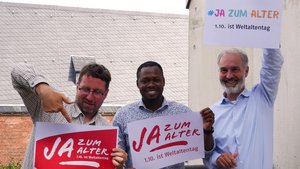 Drei Männer stehen nebeneinander. Sie halten jeweils ein Schild in der Hand mit der Aufschrift "Ja zum Alter!" Einer von ihnen zeigt mit dem Zeigefinger auf das Schild.