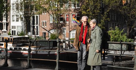 Ein älteres Pärchen steht auf einer Brücke in Amsterdam. Er sieht sie liebevoll an.