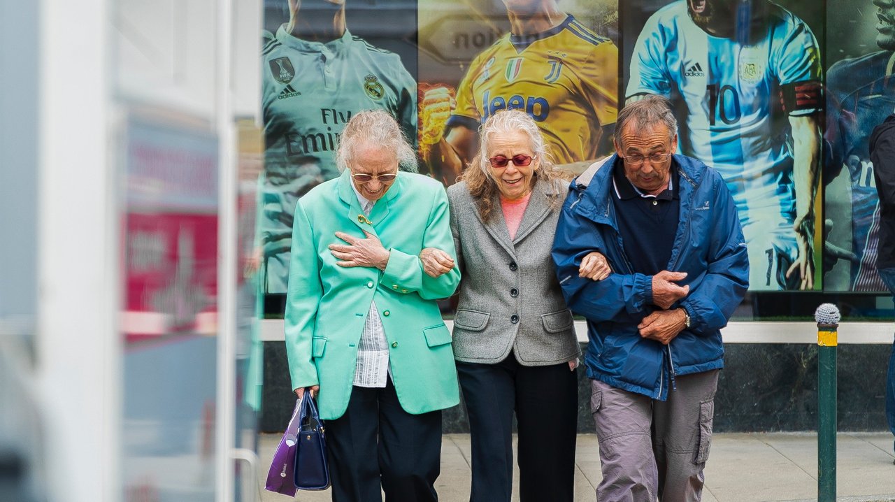 Drei ältere Menschen stehen, den Blick nach unten gerichtet, vor einem Plakat