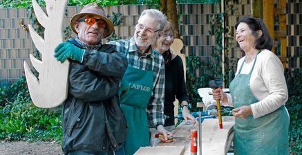 4 lachende ältere Personen stehen um einen Tisch und arbeiten mit Holz
