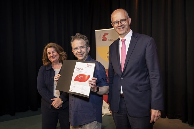 Zwei Männer, eine Frau. Der Mann, der in der Mitte steht, hält eine Urkunde in der Hand. Klick öffnet eine vergrößerte Ansicht.