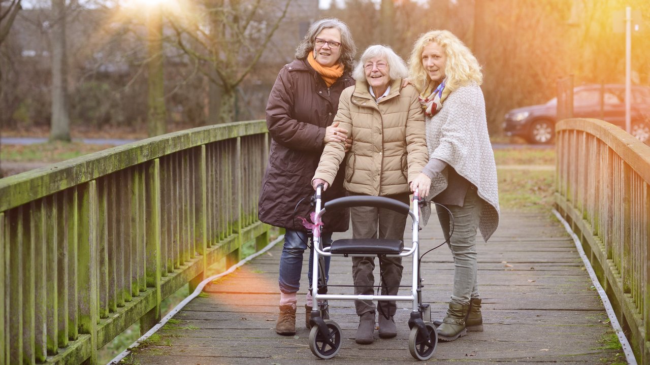 Zwei Frauen mittleren Alters und eine alte Dame mit Rollator gehen gemeinsam spazieren.