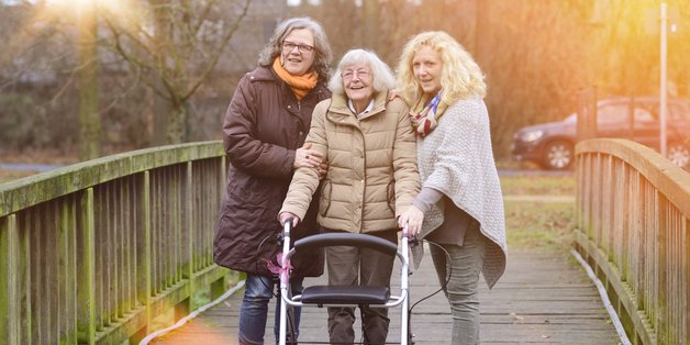 Zwei Frauen mittleren Alters und eine alte Dame mit Rollator gehen gemeinsam spazieren.