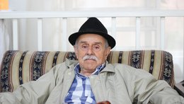 Ein alter Mann mit Hut, Jacke und Stock sitzt in einem Sessel und schaut in die Kamera.