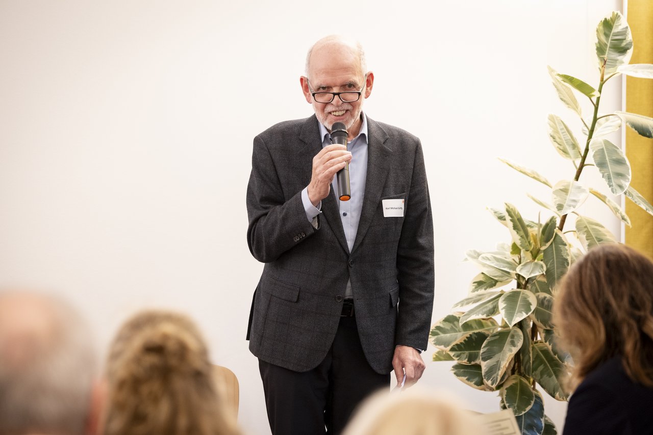 Ein älterer Mann steht vor Publikum und spricht in ein Mikrofon. Er trägt einen dunklen Anzug und darunter ein helles Hemd. Klick öffnet eine vergrößerte Ansicht.