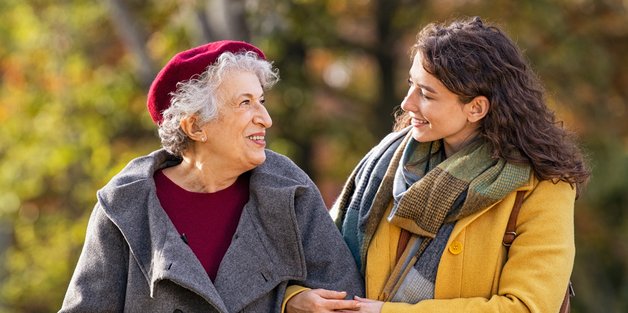 Alte und jüngere Frau machen eine Spaziergang durch einen Park. Die junge Frau hat sich bei der älteren Frau, die einen Gehstock hat, untergehakt.