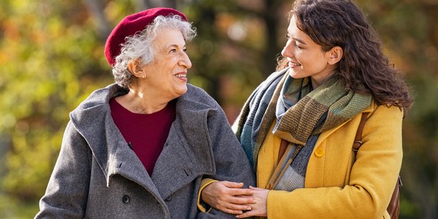 Alte und jüngere Frau machen eine Spaziergang durch einen Park. Die junge Frau hat sich bei der älteren Frau, die einen Gehstock hat, untergehakt.