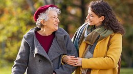 Alte und jüngere Frau machen eine Spaziergang durch einen Park. Die junge Frau hat sich bei der älteren Frau, die einen Gehstock hat, untergehakt.