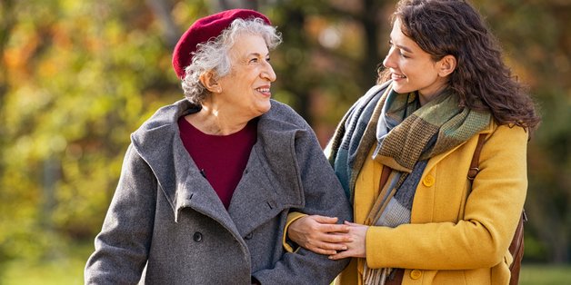 Alte und jüngere Frau machen eine Spaziergang durch einen Park. Die junge Frau hat sich bei der älteren Frau, die einen Gehstock hat, untergehakt.