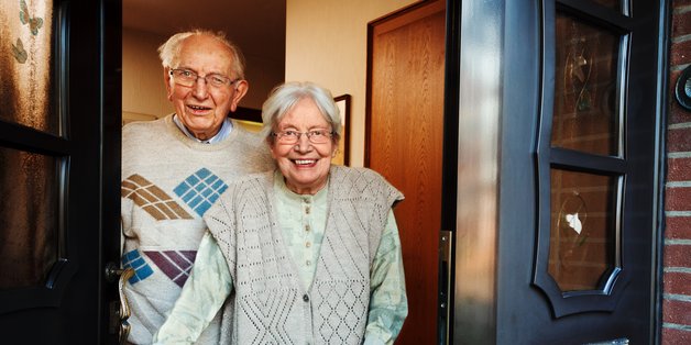 Ein alter Mann und eine alte Frau stehen in ihrer Haustür und lächeln in die Kamera.