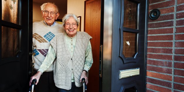 Ein alter Mann und eine alte Frau stehen in ihrer Haustür und lächeln in die Kamera.