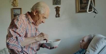 Ein älterer Mann mit einem Teller Essen steht neben einem Bett an in dem eine ältere Frau liegt. Er füttert sie.