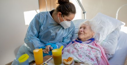 Eine junge Pflegerin in Schutzkleidung beugt sich über eine ältere Frau im Pflegebett.
