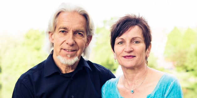 Ein älterer Mann und eine Frau sitzen auf einem Brückgeländer. Im Hintergrund sieht man Wasser und Bäume.