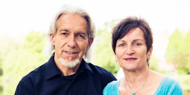 Ein älterer Mann und eine Frau sitzen auf einem Brückgeländer. Im Hintergrund sieht man Wasser und Bäume.