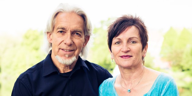 Ein älterer Mann und eine Frau sitzen auf einem Brückgeländer. Im Hintergrund sieht man Wasser und Bäume.