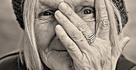 Portrait einer alten Frau mit Mütze. Sie hält sich eine Hand vor ihr Gesicht. 