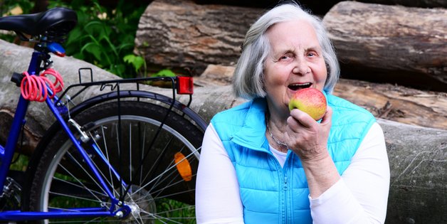 Ältere Frau isst einen Apfpel. Sie sitzt vor einem Holzstapel, vor dem ein Fahrrad abgestellt wurde.