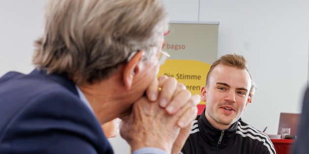 Im Tagungsraum: Junger Mann spricht zu älterem Mann, der nur von der Seite zu sehen ist.
