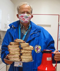 Mitarbeiter der Bahnhofsmission in blauer Dienstjacke und mit Coronamaske. In der einen Hand hält er ein Tablett mit Butterbroten. In der anderen Hand eine rote Thermoskanne.Tee