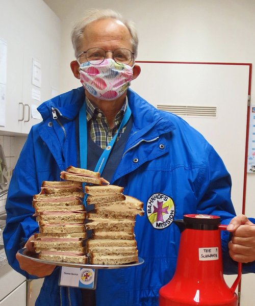 Mitarbeiter der Bahnhofsmission in blauer Dienstjacke und mit Coronamaske. In der einen Hand hält er ein Tablett mit Butterbroten. In der anderen Hand eine rote Thermoskanne.Tee