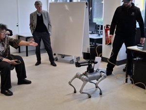 In einem großen Büroraum wurde in der Mitte ein freier Platz geschaffen. Dort stehen drei Personen um einen Roboter, der wie ein vierbeiniges Tier dargestellt wird. Er hält in einer Greifvorrichtung ein Buch.