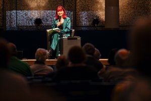 Auf der Bühne: Eine junge Frau sitzt in einem schwarzen Sessel. Sie hat ein aufgeschlagenes Buch in der Hand und spricht in ein Mikrofon. Sie trägt einen Hosenanzug in aufälligem Grün und hat rotes schulterlanges Haar.