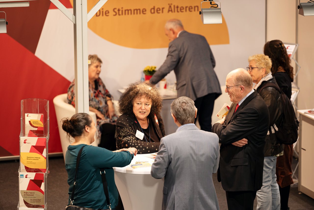Am BAGSO-Stand stehen um einen Stehtisch mehrere Personen. Im Mittelpunkt die BAGSO-Vorsitzende Regina Görner, ein ältere Frau mit dunklem krausen Haar, dunkel gekleidet. Sie spricht gerade in die Runde. Klick öffnet eine vergrößerte Ansicht.