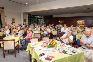 Ein großer Saal mit Tischen auf den grüne Tischdecken liegen, darauf liegen Unterlagen und stehen Getränke. Um die Tische herum sitzen Menschen, die nach vorne schauen und klatschen.