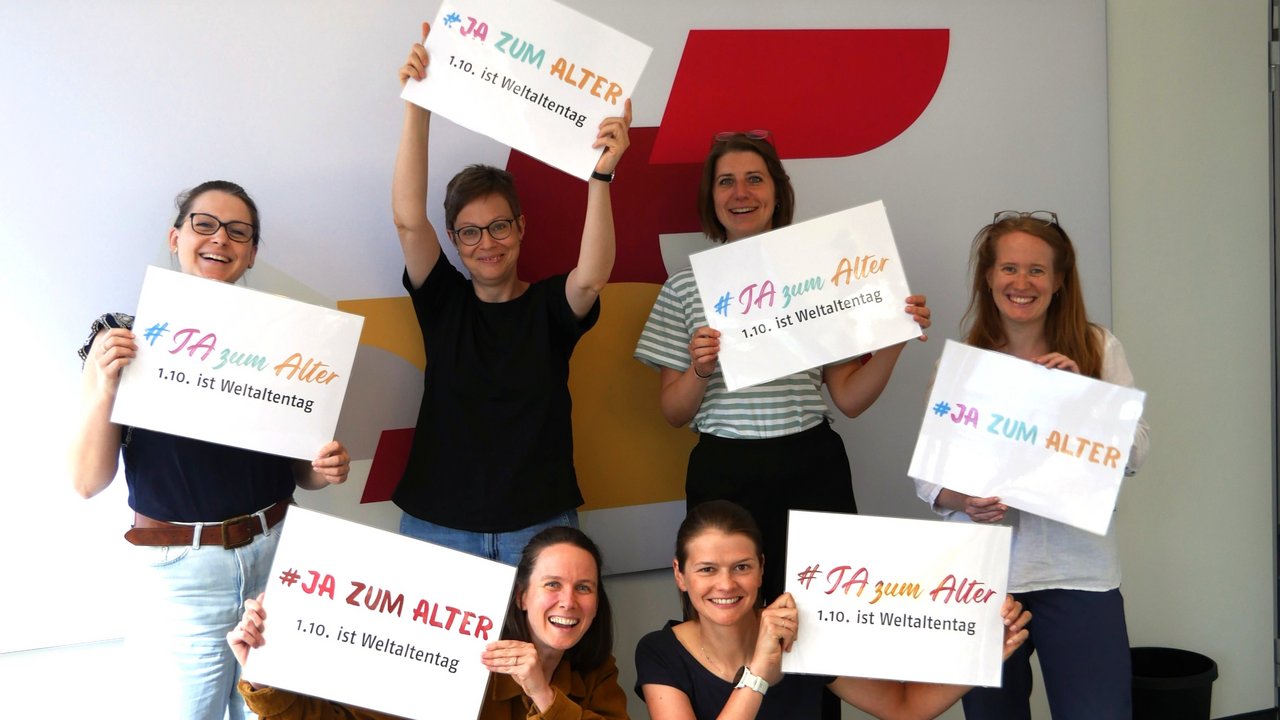 Sechs Frauen stehen vor einer Fotowand, die BAGSO-Logo-Elemente zeigt. Sie lachen alle und halten jeweils ein Schild in der Hand mit der Aufschrift "Ja zum Alter!" Klick öffnet eine vergrößerte Ansicht.