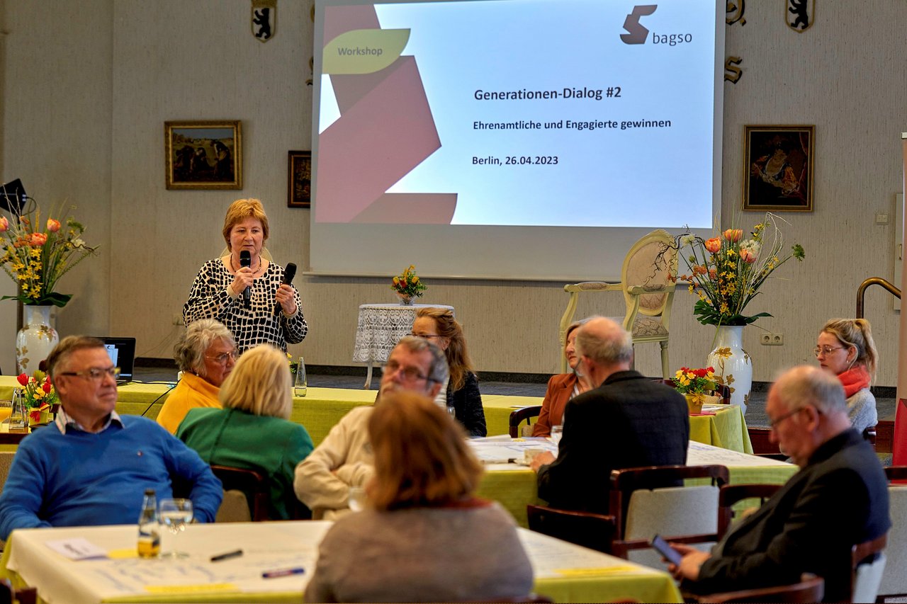 Eine Frau mittleren Alters hält zwei Mikrofone in der Hand. In eine Mikrofon spricht sie gerade zu den Teilnehmern, die in einem Saal an Tischen sitzen. Im Hintergrund ist eine Leinwand mit einer Powerpoint zu sehen.