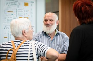 Ein alter Mann mit grauem Vollbart und Halbglatze. Er trägt ein blau gstreiftes Hemd und schaut mit den Augen lächelnd nach links. Ihm gegenüber stehen zwei ältere Frauen mit dem Rücken zur Kamera.