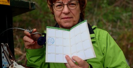 Ältere Frau in grüner Wanderjacke stempelt ein Wanderheft ab