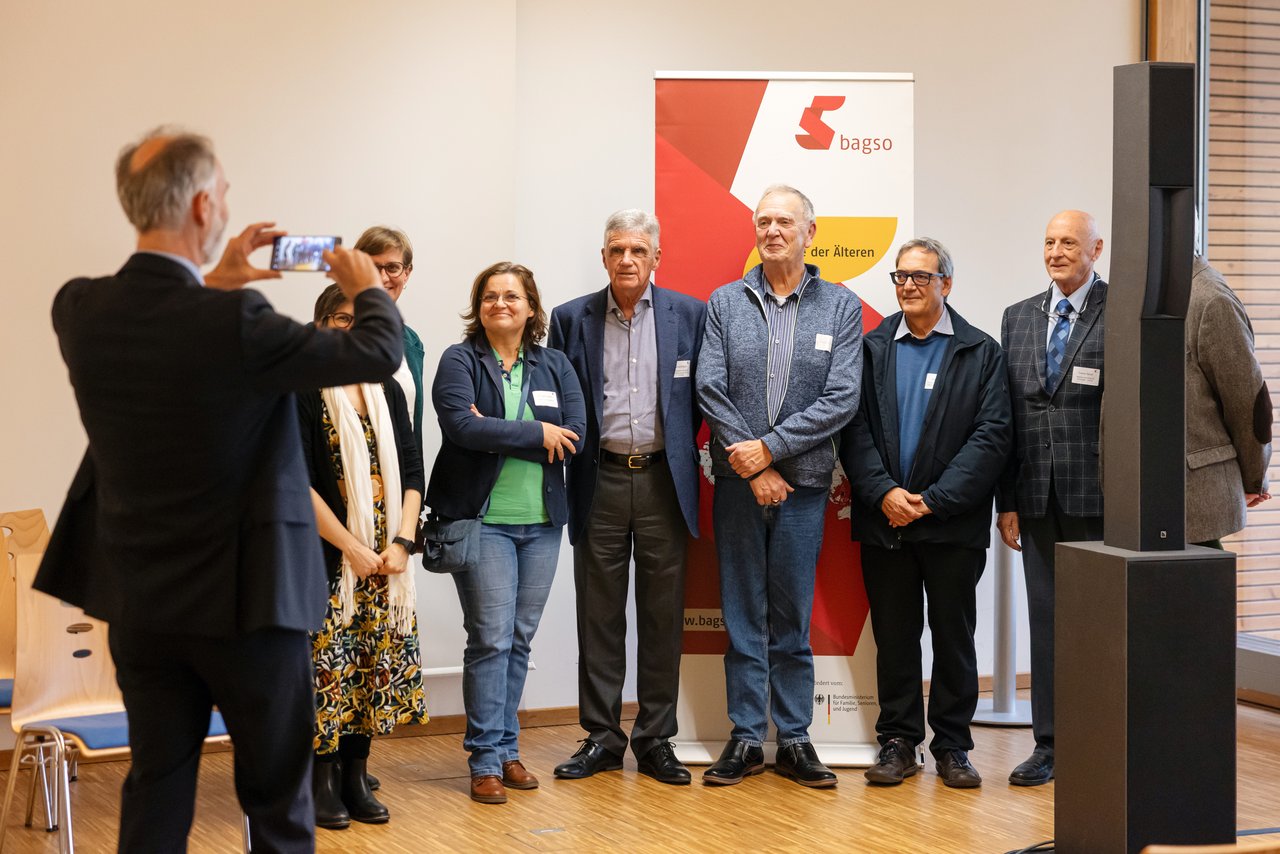 Ein Mann mittleren Alters fotografiert eine Gruppe von Personen unterschiedlichen Alters vor einem Banner mit der Aufschrift BAGSO.
