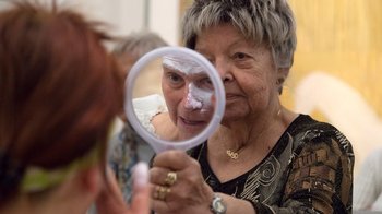 Zwei Frauen stehen sich gegenüber und schauen jeweils in einen Handspiegel. In einem Spiegel sieht man ein weiß geschminktes Gesicht.