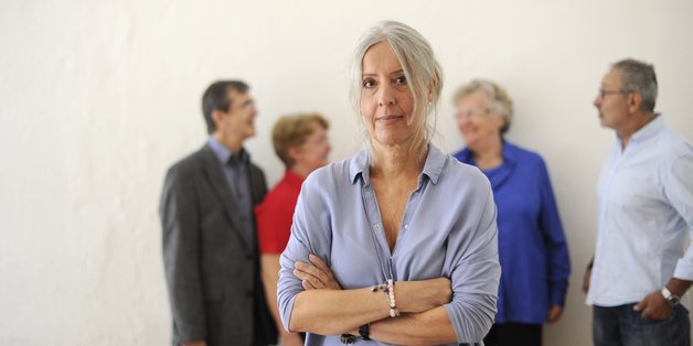 Frau steht allein mit verschränkten Armen im Vordergrund. Gruppe miteinander sprechender Menschen im Hintergrund