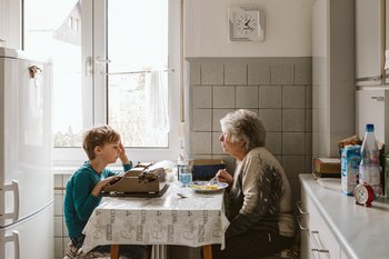 Ältere Frau sitzt am Küchentisch und isst. Ihr Enkel gegenüber schreibt auf einer alten Schreibmaschine.