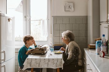 ©Julia Otto Ältere Frau sitzt am Küchentisch und isst. Ihr Enkel gegenüber schreibt auf einer alten Schreibmaschine.