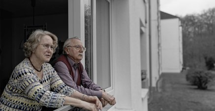 Ein älteres Ehepaar steht auf dem Balkon einer recht tristen Wohnanlage und schaut gemeinhoffnungsvoll in die Ferne