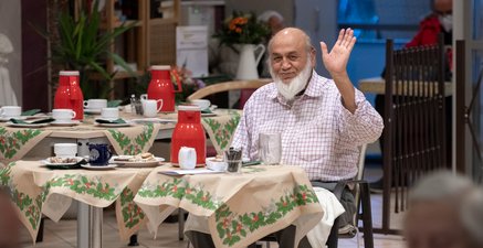 Lächelnder, dunkelhäutiger Mann mit Halbglatze und weißem Bart sitzt im leeren Festsaal, er winkt