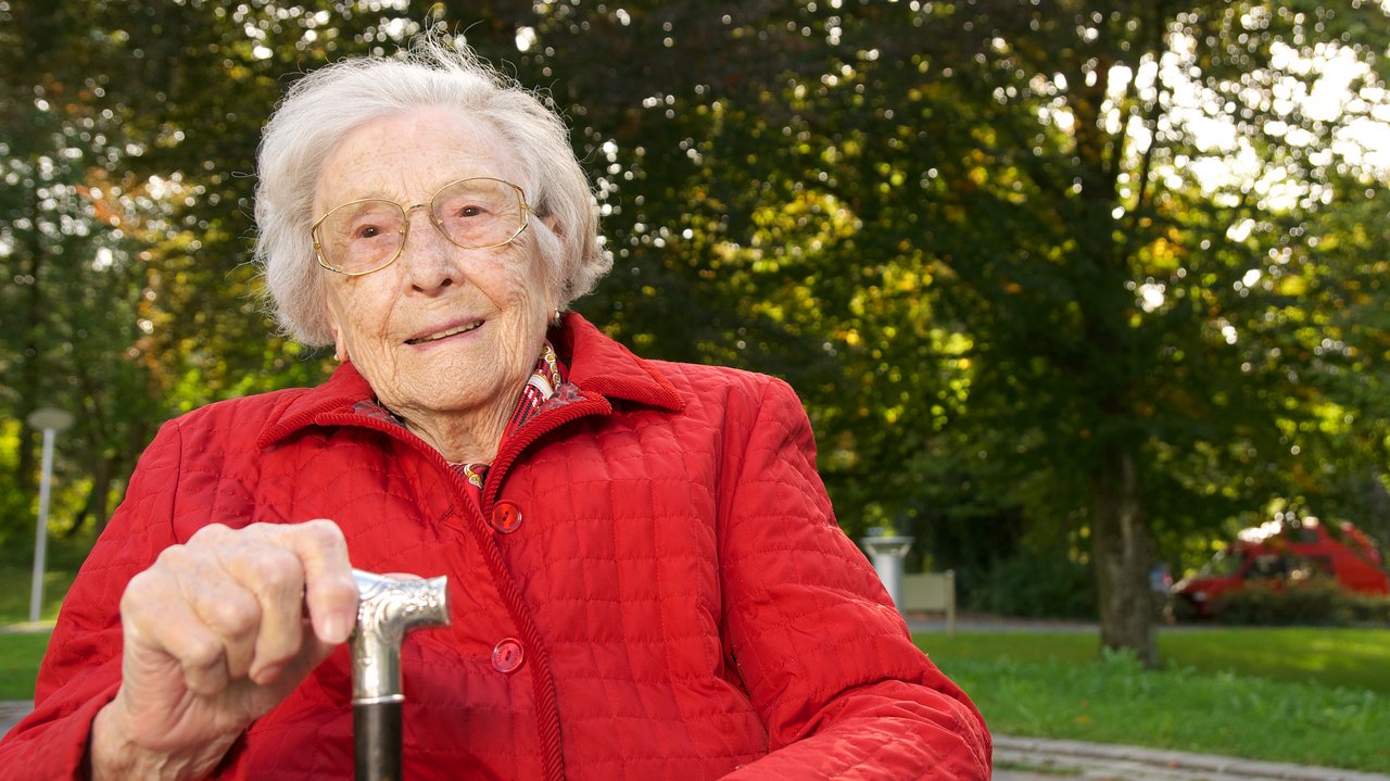 Alte Frau mit Stock sitzt auf einer Bank.