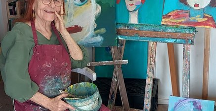 Ältere Malerin sitzt in ihrem Atelier. Hinter ihr unzählige farbenfrohe Portraitbilder.