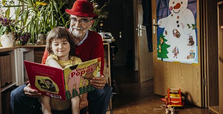 Ein weißhaariger Mann und ein fünfjähriges Mädchen sitzen auf einer Bank in einer Wohnung und lesen interessiert.