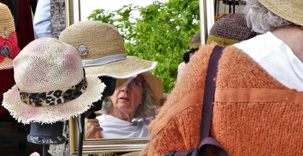 Seniorin hat in einem Hutladen einen Hut ausgewählt und begutachtet sich im Spiegel.